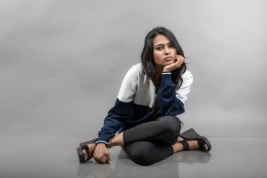 Gri arka planda günlük kışlık giysili Hintli genç bayan model. Siyah tayt giyen uzun siyah saçlı bir manken, mavi beyaz bir kazak..