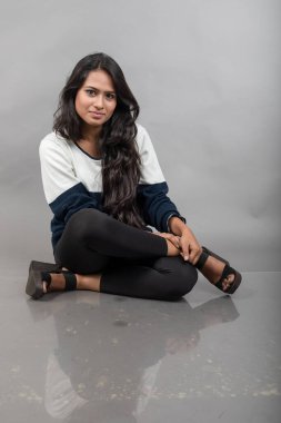 Gri arka planda günlük kışlık giysili Hintli genç bayan model. Siyah tayt giyen uzun siyah saçlı bir manken, mavi beyaz bir kazak..