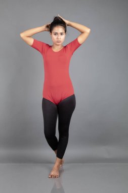 Kırmızı tişörtlü, siyah taytlı, yoga ve esneme egzersizleri yapan sağlıklı genç bir kadın gri arka planda izole edilmiş.