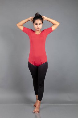 Kırmızı tişörtlü, siyah taytlı, yoga ve esneme egzersizleri yapan sağlıklı genç bir kadın gri arka planda izole edilmiş.