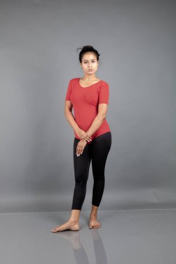 Kırmızı tişörtlü, siyah taytlı, yoga ve esneme egzersizleri yapan sağlıklı genç bir kadın gri arka planda izole edilmiş.