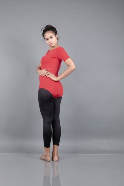 Kırmızı tişörtlü, siyah taytlı, yoga ve esneme egzersizleri yapan sağlıklı genç bir kadın gri arka planda izole edilmiş.