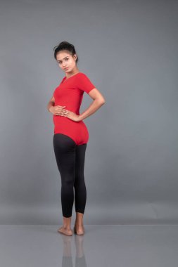 Kırmızı tişörtlü, siyah taytlı, yoga ve esneme egzersizleri yapan sağlıklı genç bir kadın gri arka planda izole edilmiş.