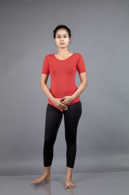 Kırmızı tişörtlü, siyah taytlı, yoga ve esneme egzersizleri yapan sağlıklı genç bir kadın gri arka planda izole edilmiş.