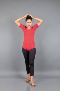 Kırmızı tişörtlü, siyah taytlı, yoga ve esneme egzersizleri yapan sağlıklı genç bir kadın gri arka planda izole edilmiş.