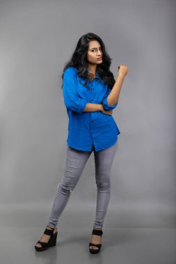 Gri arka plan karşısında gündelik giyimli Hintli genç manken. Stok fotoğrafı. Uzun siyah saç modeli mavi gömlek giyiyor..