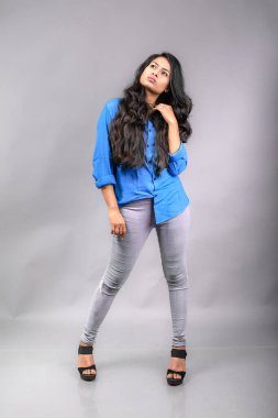 Gri arka plan karşısında gündelik giyimli Hintli genç manken. Stok fotoğrafı. Uzun siyah saç modeli mavi gömlek giyiyor..