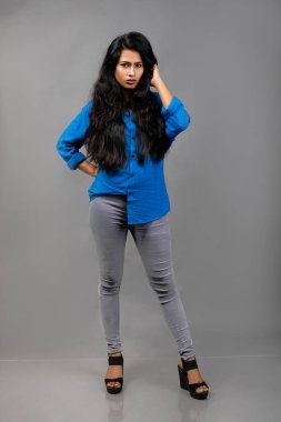 Gri arka plan karşısında gündelik giyimli Hintli genç manken. Stok fotoğrafı. Uzun siyah saç modeli mavi gömlek giyiyor..