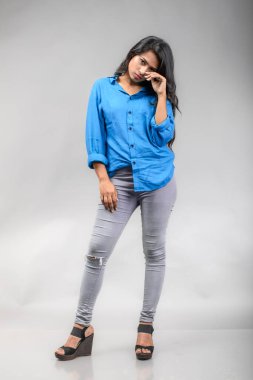 Gri arka plan karşısında gündelik giyimli Hintli genç manken. Stok fotoğrafı. Uzun siyah saç modeli mavi gömlek giyiyor..