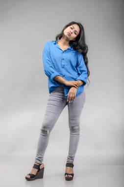 Gri arka plan karşısında gündelik giyimli Hintli genç manken. Stok fotoğrafı. Uzun siyah saç modeli mavi gömlek giyiyor..