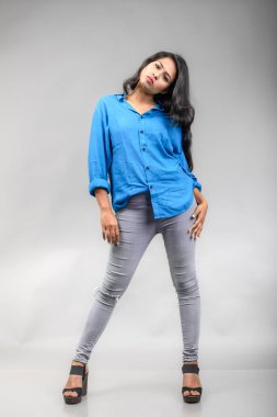 Gri arka plan karşısında gündelik giyimli Hintli genç manken. Stok fotoğrafı. Uzun siyah saç modeli mavi gömlek giyiyor..