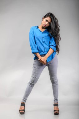 Gri arka plan karşısında gündelik giyimli Hintli genç manken. Stok fotoğrafı. Uzun siyah saç modeli mavi gömlek giyiyor..