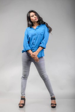 Gri arka plan karşısında gündelik giyimli Hintli genç manken. Stok fotoğrafı. Uzun siyah saç modeli mavi gömlek giyiyor..