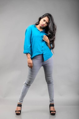 Gri arka plan karşısında gündelik giyimli Hintli genç manken. Stok fotoğrafı. Uzun siyah saç modeli mavi gömlek giyiyor..