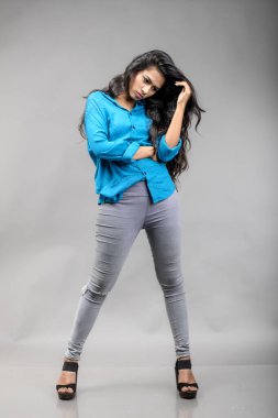 Gri arka plan karşısında gündelik giyimli Hintli genç manken. Stok fotoğrafı. Uzun siyah saç modeli mavi gömlek giyiyor..