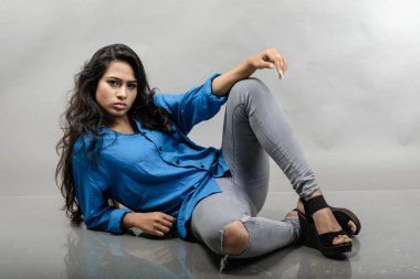 Gri arka plan karşısında gündelik giyimli Hintli genç manken. Stok fotoğrafı. Uzun siyah saç modeli mavi gömlek, gri kot pantolon ve siyah topuklu ayakkabı giyiyor..