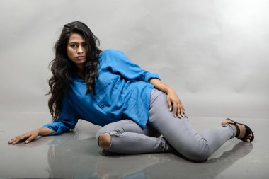 Gri arka plan karşısında gündelik giyimli Hintli genç manken. Stok fotoğrafı. Uzun siyah saç modeli mavi gömlek, gri kot pantolon ve siyah topuklu ayakkabı giyiyor..