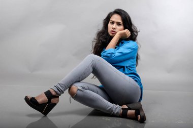 Gri arka plan karşısında gündelik giyimli Hintli genç manken. Stok fotoğrafı. Uzun siyah saç modeli mavi gömlek, gri kot pantolon ve siyah topuklu ayakkabı giyiyor..