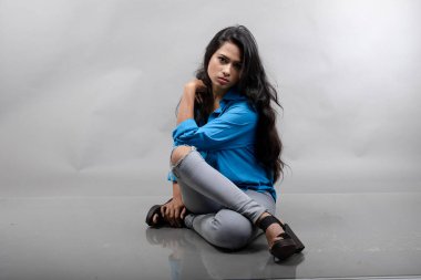 Gri arka plan karşısında gündelik giyimli Hintli genç manken. Stok fotoğrafı. Uzun siyah saç modeli mavi gömlek, gri kot pantolon ve siyah topuklu ayakkabı giyiyor..
