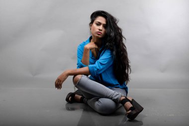 Gri arka plan karşısında gündelik giyimli Hintli genç manken. Stok fotoğrafı. Uzun siyah saç modeli mavi gömlek, gri kot pantolon ve siyah topuklu ayakkabı giyiyor..