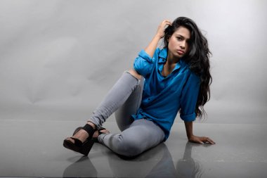 Gri arka plan karşısında gündelik giyimli Hintli genç manken. Stok fotoğrafı. Uzun siyah saç modeli mavi gömlek, gri kot pantolon ve siyah topuklu ayakkabı giyiyor..