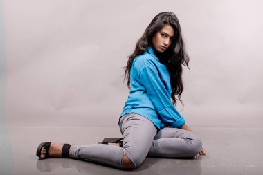 Gri arka plan karşısında gündelik giyimli Hintli genç manken. Stok fotoğrafı. Uzun siyah saç modeli mavi gömlek, gri kot pantolon ve siyah topuklu ayakkabı giyiyor..