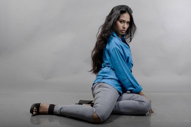 Gri arka plan karşısında gündelik giyimli Hintli genç manken. Stok fotoğrafı. Uzun siyah saç modeli mavi gömlek, gri kot pantolon ve siyah topuklu ayakkabı giyiyor..