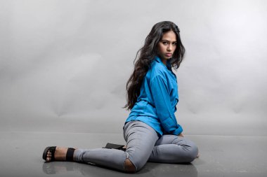 Gri arka plan karşısında gündelik giyimli Hintli genç manken. Stok fotoğrafı. Uzun siyah saç modeli mavi gömlek, gri kot pantolon ve siyah topuklu ayakkabı giyiyor..