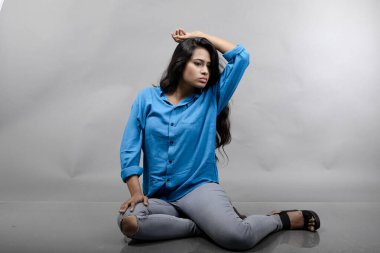 Gri arka plan karşısında gündelik giyimli Hintli genç manken. Stok fotoğrafı. Uzun siyah saç modeli mavi gömlek, gri kot pantolon ve siyah topuklu ayakkabı giyiyor..