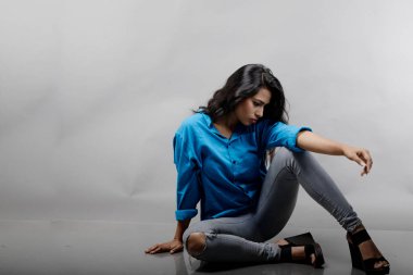 Gri arka plan karşısında gündelik giyimli Hintli genç manken. Stok fotoğrafı. Uzun siyah saç modeli mavi gömlek, gri kot pantolon ve siyah topuklu ayakkabı giyiyor..