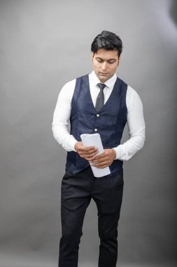 Hintli genç iş adamı resmi bir klasör üzerinde sunum yapıyor. Beyaz gömlek, siyah kravat ve gri arka planda siyah ceket giyiyor. Erkek modeli.