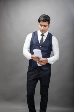 Hintli genç iş adamı resmi bir klasör üzerinde sunum yapıyor. Beyaz gömlek, siyah kravat ve gri arka planda siyah ceket giyiyor. Erkek modeli.