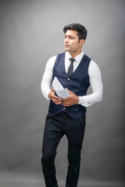 Hintli genç iş adamı resmi bir klasör üzerinde sunum yapıyor. Beyaz gömlek, siyah kravat ve gri arka planda siyah ceket giyiyor. Erkek modeli.