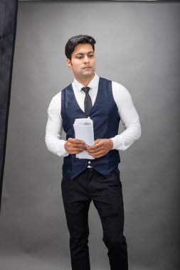 Hintli genç iş adamı resmi bir klasör üzerinde sunum yapıyor. Beyaz gömlek, siyah kravat ve gri arka planda siyah ceket giyiyor. Erkek modeli.