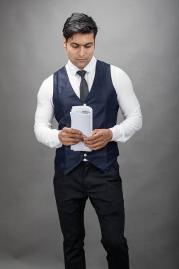 Hintli genç iş adamı resmi bir klasör üzerinde sunum yapıyor. Beyaz gömlek, siyah kravat ve gri arka planda siyah ceket giyiyor. Erkek modeli.