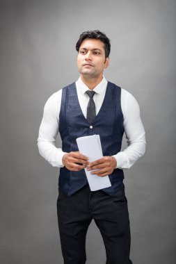 Hintli genç iş adamı resmi bir klasör üzerinde sunum yapıyor. Beyaz gömlek, siyah kravat ve gri arka planda siyah ceket giyiyor. Erkek modeli.