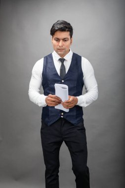 Hintli genç iş adamı resmi bir klasör üzerinde sunum yapıyor. Beyaz gömlek, siyah kravat ve gri arka planda siyah ceket giyiyor. Erkek modeli.