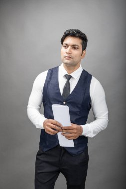 Hintli genç iş adamı resmi bir klasör üzerinde sunum yapıyor. Beyaz gömlek, siyah kravat ve gri arka planda siyah ceket giyiyor. Erkek modeli.
