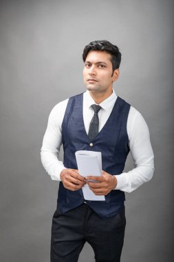 Hintli genç iş adamı resmi bir klasör üzerinde sunum yapıyor. Beyaz gömlek, siyah kravat ve gri arka planda siyah ceket giyiyor. Erkek modeli.