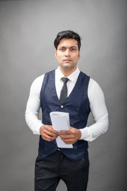 Hintli genç iş adamı resmi bir klasör üzerinde sunum yapıyor. Beyaz gömlek, siyah kravat ve gri arka planda siyah ceket giyiyor. Erkek modeli.
