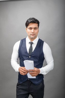 Hintli genç iş adamı resmi bir klasör üzerinde sunum yapıyor. Beyaz gömlek, siyah kravat ve gri arka planda siyah ceket giyiyor. Erkek model. Hintli genç iş adamı bir konu tutucu klasöründe sunum yapıyor