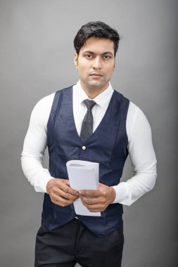 Hintli genç iş adamı resmi bir klasör üzerinde sunum yapıyor. Beyaz gömlek, siyah kravat ve gri arka planda siyah ceket giyiyor. Erkek modeli.