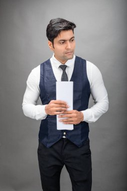 Hintli genç iş adamı resmi bir klasör üzerinde sunum yapıyor. Beyaz gömlek, siyah kravat ve gri arka planda siyah ceket giyiyor. Erkek modeli.