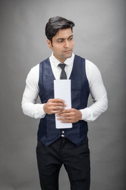 Hintli genç iş adamı resmi bir klasör üzerinde sunum yapıyor. Beyaz gömlek, siyah kravat ve gri arka planda siyah ceket giyiyor. Erkek modeli.