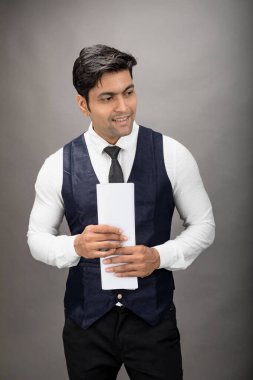 Hintli genç iş adamı resmi bir klasör üzerinde sunum yapıyor. Beyaz gömlek, siyah kravat ve gri arka planda siyah ceket giyiyor. Erkek modeli.
