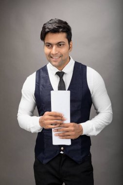 Hintli genç iş adamı resmi bir klasör üzerinde sunum yapıyor. Beyaz gömlek, siyah kravat ve gri arka planda siyah ceket giyiyor. Erkek modeli.