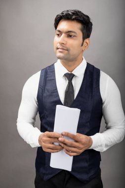 Hintli genç iş adamı resmi bir klasör üzerinde sunum yapıyor. Beyaz gömlek, siyah kravat ve gri arka planda siyah ceket giyiyor. Erkek modeli.