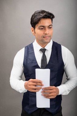 Hintli genç iş adamı resmi bir klasör üzerinde sunum yapıyor. Beyaz gömlek, siyah kravat ve gri arka planda siyah ceket giyiyor. Erkek modeli.
