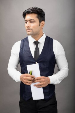 Hintli genç iş adamı resmi bir klasör üzerinde sunum yapıyor. Beyaz gömlek, siyah kravat ve gri arka planda siyah ceket giyiyor. Erkek modeli.