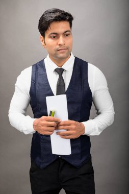 Hintli genç iş adamı resmi bir klasör üzerinde sunum yapıyor. Beyaz gömlek, siyah kravat ve gri arka planda siyah ceket giyiyor. Erkek modeli.